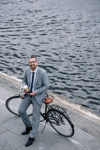 コーヒーとスマート フォン立って岸壁に自転車を持ったビジネスマン  — 無料ストックフォト