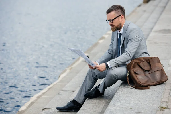 handsome businessman in grey suit with leather bag reading newspaper on quay