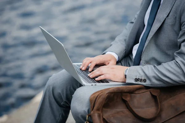 cropped view of businessman using laptop and sitting on quay with leather bag