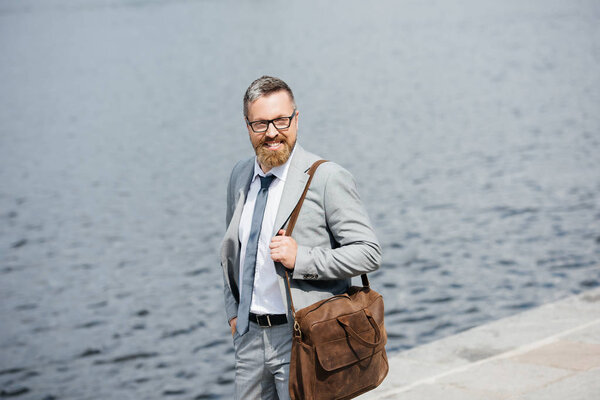 handsome businessman with leather bag walking on quay near river 