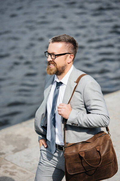 handsome bearded businessman in grey suit with leather bag walking on quay  