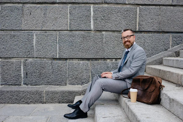businessman with bag and disposable cup of coffee using laptop and sitting on stairs