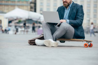 dizüstü bilgisayar ile kulaklık kullanarak ve longboard şehirde oturan adam görünümünü kırpılmış