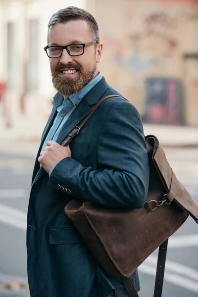 portrait of stylish cheerful bearded man with leather bag walking in city