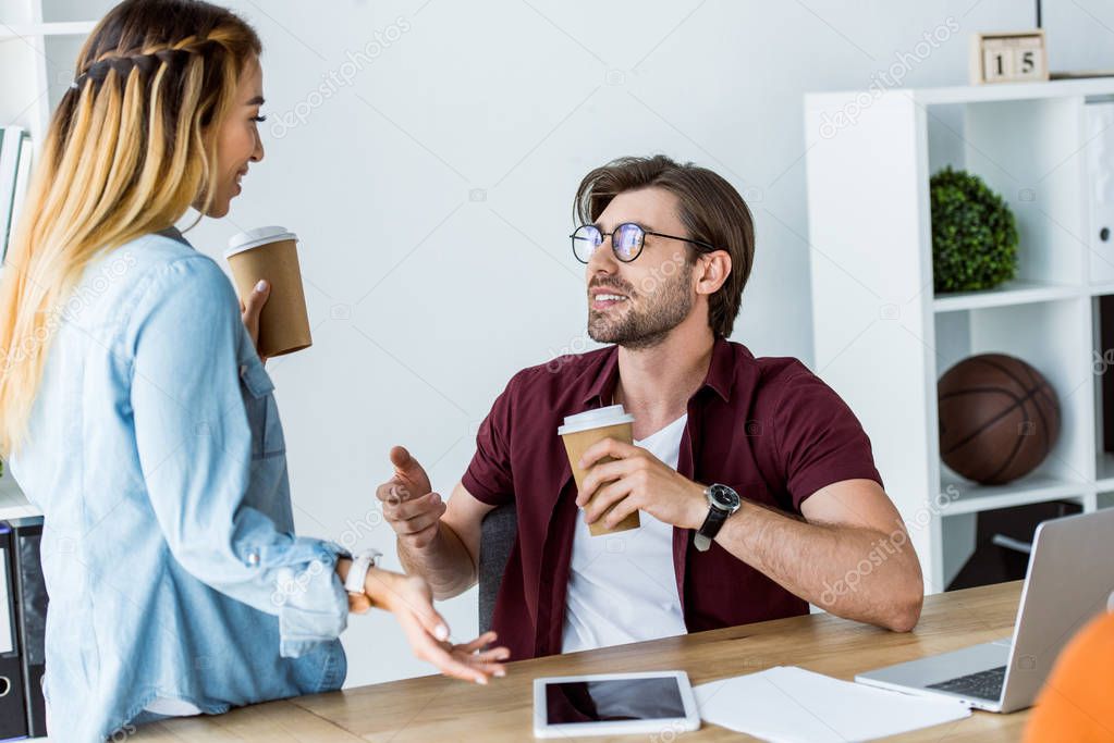 multicultural colleagues talking and holding disposable coffee cups in office