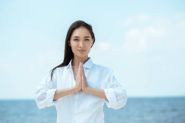 focused asian woman doing namaste mudra gesture in front of sea  clipart