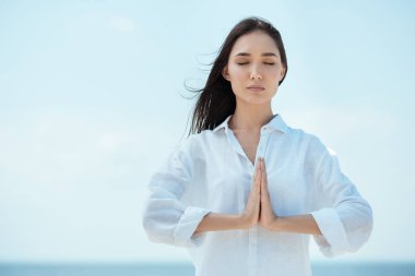 concentrated asian woman with closed eyes doing namaste mudra gesture in front of sea  clipart