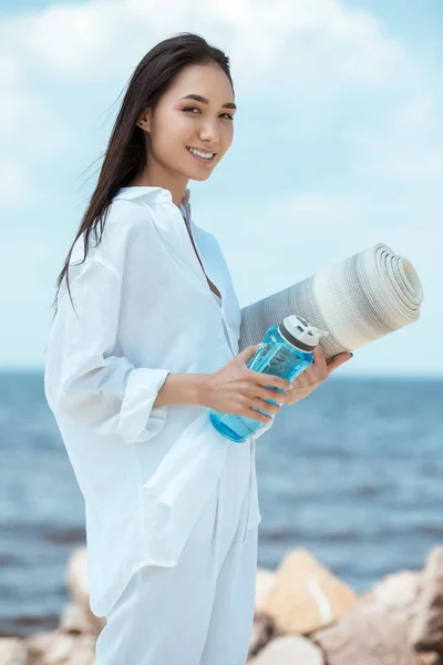 ビーチで水とヨガのマットのスポーツ ボトルを保持しているアジアの若い女性を笑顔 — ストック写真