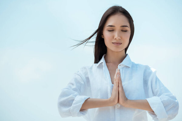 asian woman with closed eyes doing namaste mudra gesture against blue sky