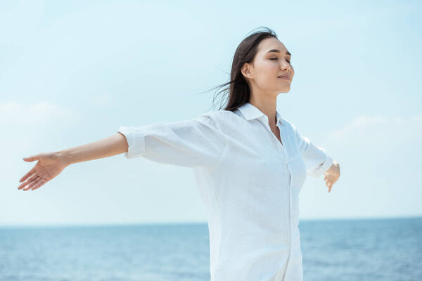 young asian woman with closed eyes standing with arms outstretched by sea 
