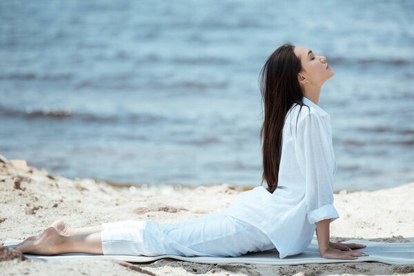 side view of focused asian woman practicing yoga on mat by sea 