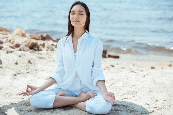 asian woman with closed eyes meditating with akash mudra gesture in lotus position on beach 