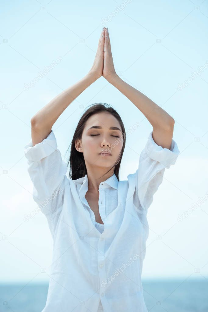 young asian woman with closed eyes standing in asana vrikshasana (tree pose) by sea