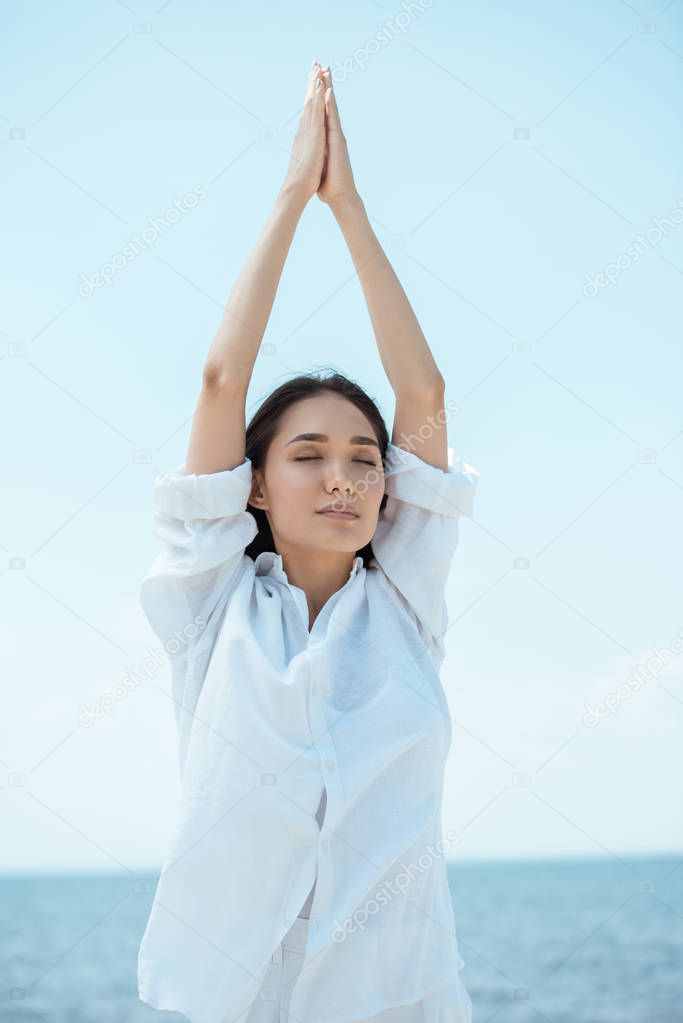  concentrated asian woman with closed eyes standing in asana vrikshasana (tree pose) by sea