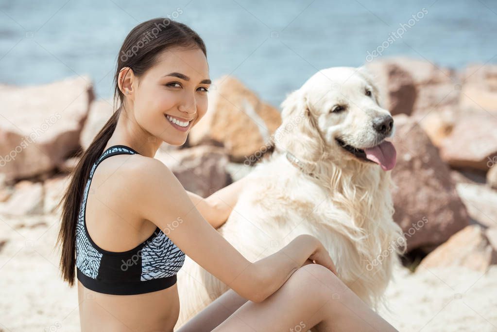 selective focus of smiling asian woman sitting with golden retriever on beach 