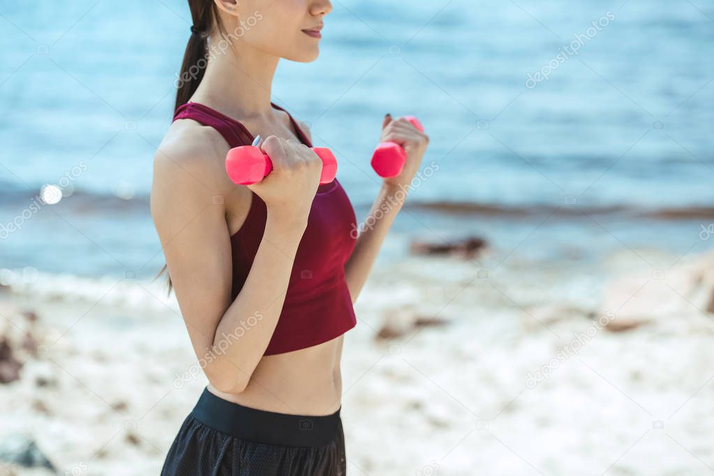 partial view of asian sportswoman exercising with dumbbells on beach