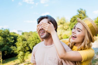 side view of happy woman covering boyfriends eyes in park on summer day clipart