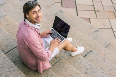 adam adımları sokakta oturan laptop ve boyun kulaklık ile gülümseyen, yüksek açılı görünüş