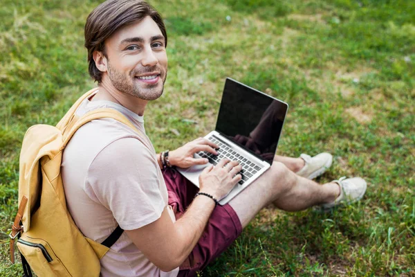 side view of smiling man with backpack and laptop sitting on green grass in park