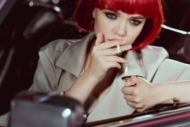 Sigara ve arabanın içinde otururken kameraya bakarak kırmızı peruk çekici kız 