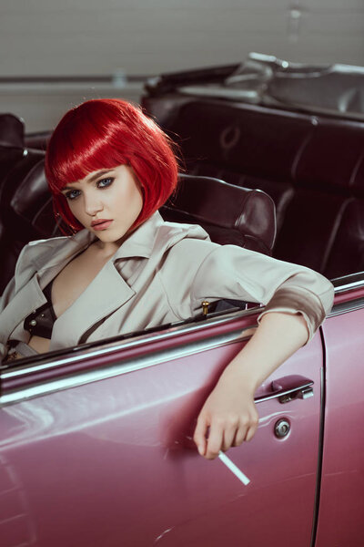 beautiful young woman in red wig holding cigarette and sitting in vintage car 