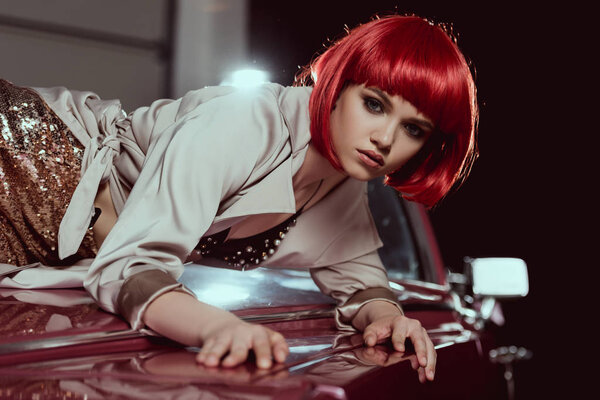 beautiful young woman in red wig and stylish trench coat looking at camera while lying at car   