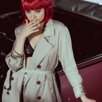 Hermosa mujer en peluca roja y gabardina fumar cigarrillo y mirando a la cámara cerca del coche