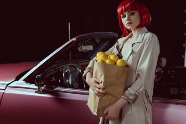 kadın kırmızı peruk ve şık limon ile kağıt torba tutarak ve kamera retro car yakınındaki dururken bakarak trençkot  