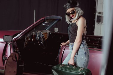 stylish blonde girl in sunglases and beret holding retro suitcase and looking at camera while standing near maroon car  clipart