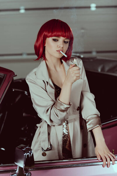 beautiful young woman in red wig and stylish trench coat smoking cigarette in classic car 