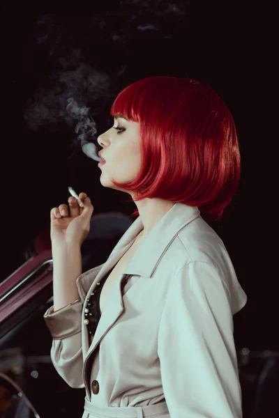美丽的女孩在红色假发和时尚的沟槽大衣吸烟香烟在汽车侧面视图 — 图库照片