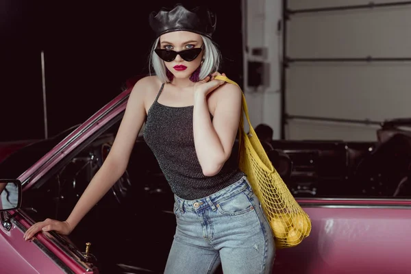 レモンとひも付きの袋を持って 車の近くに立っている間カメラ目線はベレー帽とサングラスでスタイリッシュなブロンドの女の子 — ストック写真