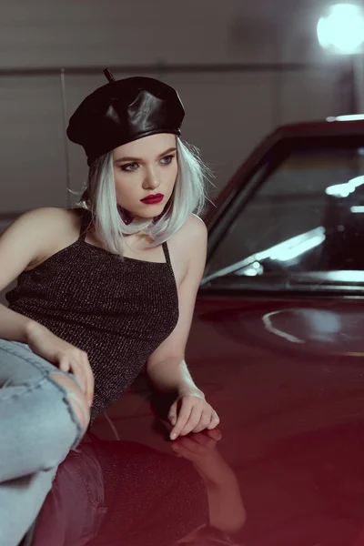 beautiful blonde woman in black beret lying on maroon car and looking away