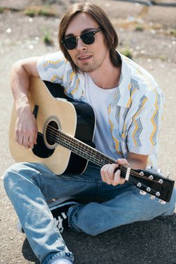 Young man playing guitar and sitting on ground in urban environment clipart