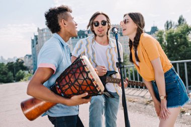 Team of young friends with musical instruments singing by microphone in urban environment clipart