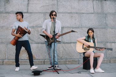 Young and happy street musicians band with guitars and djembe in city clipart