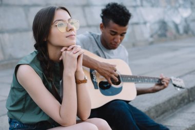 Dreamy girl listening to African american man playing guitar in city