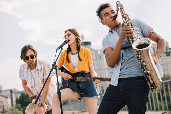 Multiracial young people with guitar, djembe and saxophone performing on street