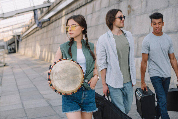 Team of young male and female friends walking and carrying musical instruments in urban environment