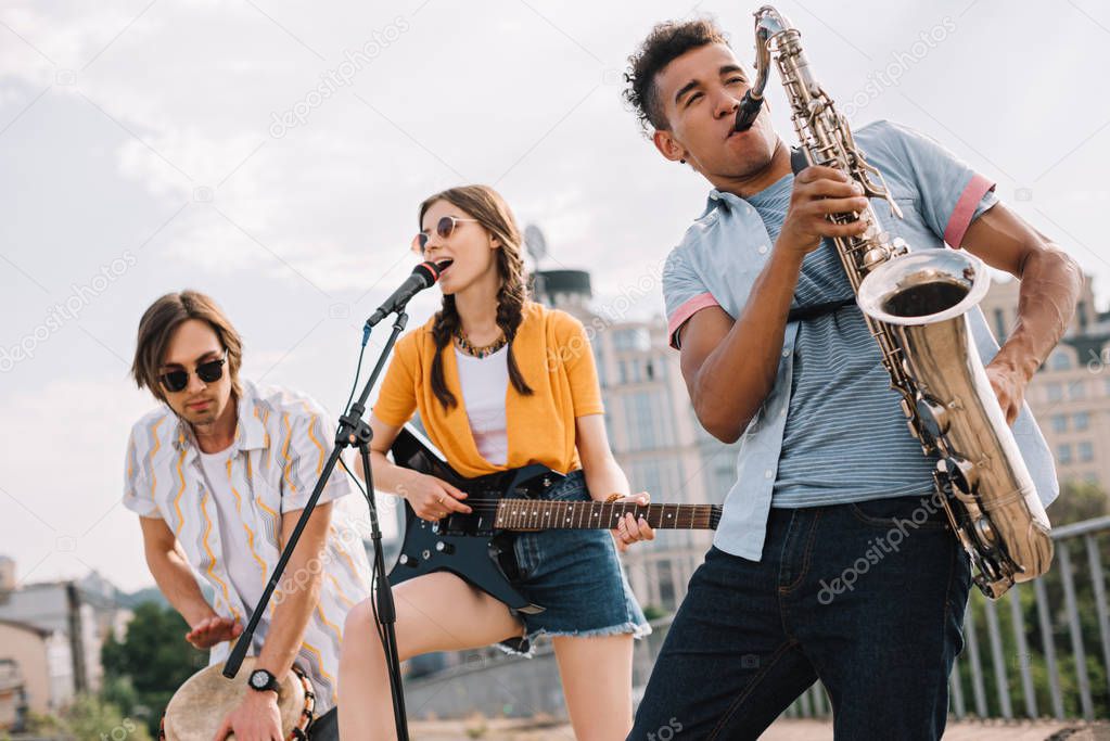 Multiracial young people with guitar, djembe and saxophone performing on street