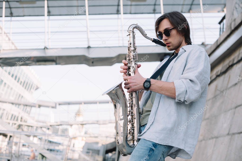 Young man in sunglasses adjusting saxophone on sunny city street