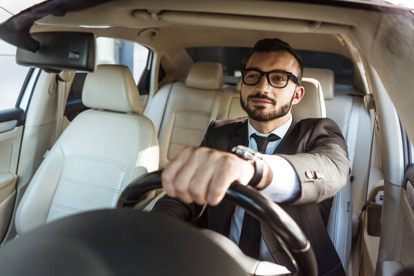 cheerful handsome driver in suit and glasses driving car