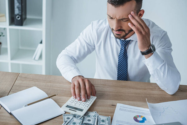 задумчивый финансист считает деньги с калькулятором в офисе и с головной болью
