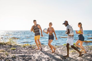 mutlu genç çok ırklı arkadaşlarıyla top ve birlikte kumsalda yürüyen sırt çantaları