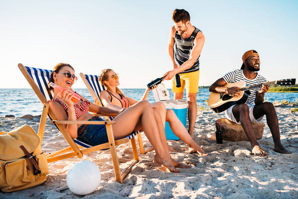 happy young multiethnic friends with guitar and drinks spending time together on sandy beach 