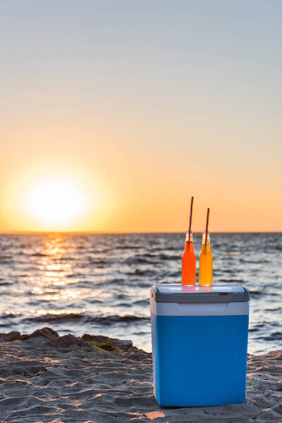 glass bottles with summer beverages and straws on cooler at sandy beach at sunset