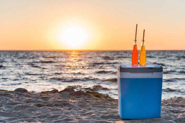 glass bottles with summer drinks and straws on cooler at sandy beach 