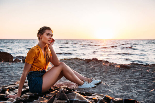 pensive young woman sitting on plaid at beautiful sea coast at sunset 