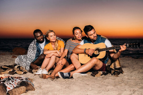 happy young multiethnic friends enjoying guitar on beach at sunset