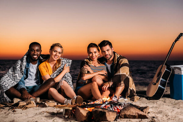 happy young people smiling at camera while sitting near bonfire on sandy beach at sunset 